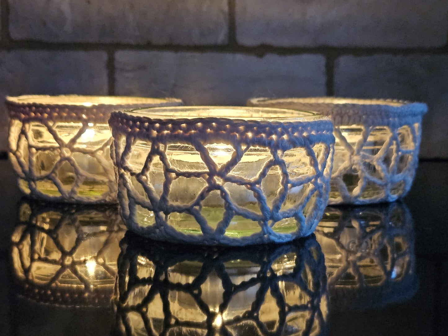 Three glass candle holders with lace-like crochet patterns on a reflective surface.