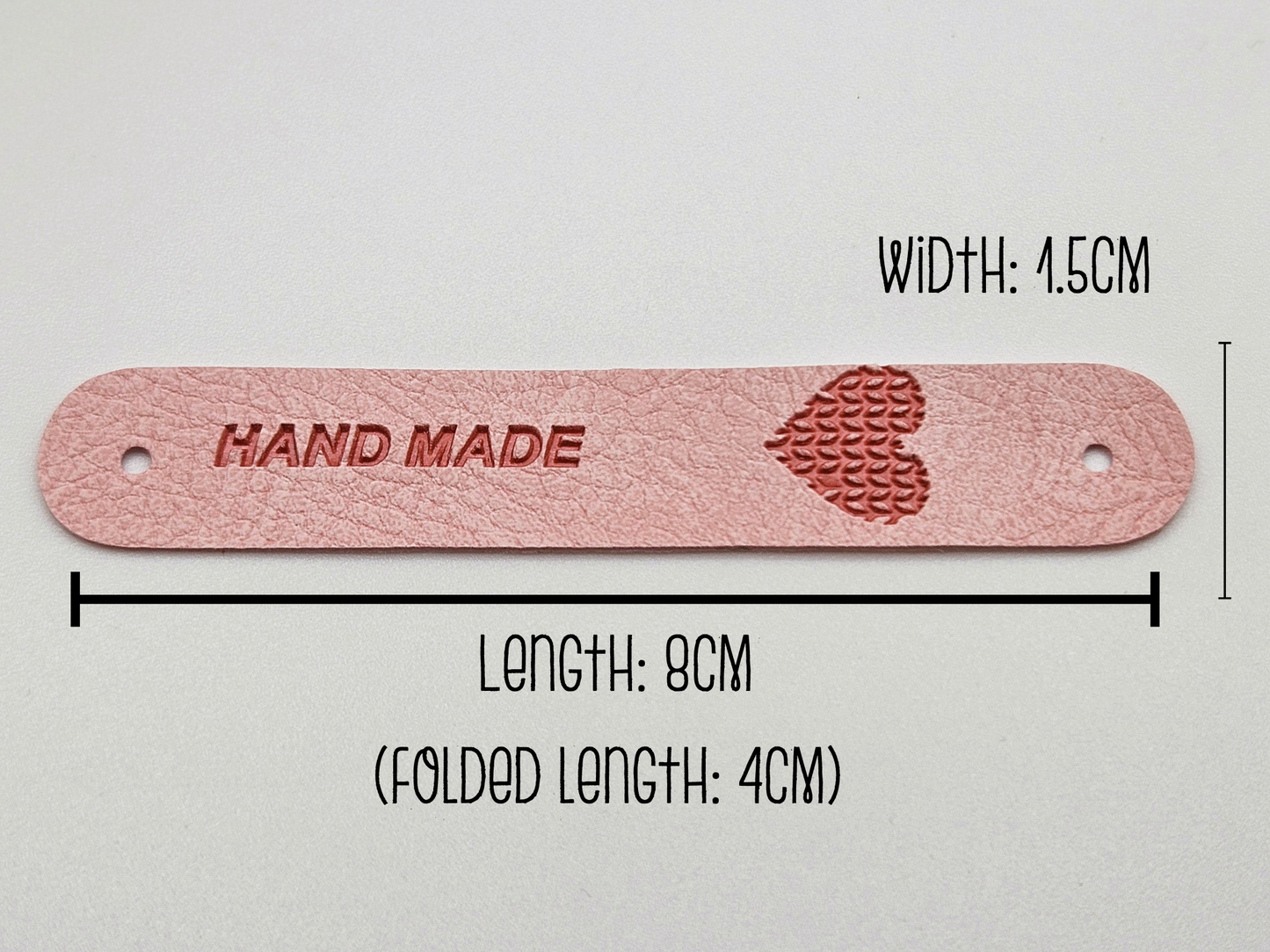 A close-up of a pink faux leather hand made tag with measurements displayed on the image, showing the heart design and snap-on popper. Measurements indicate width is 1.5cm and length 8cm. Folded length is 4cm.