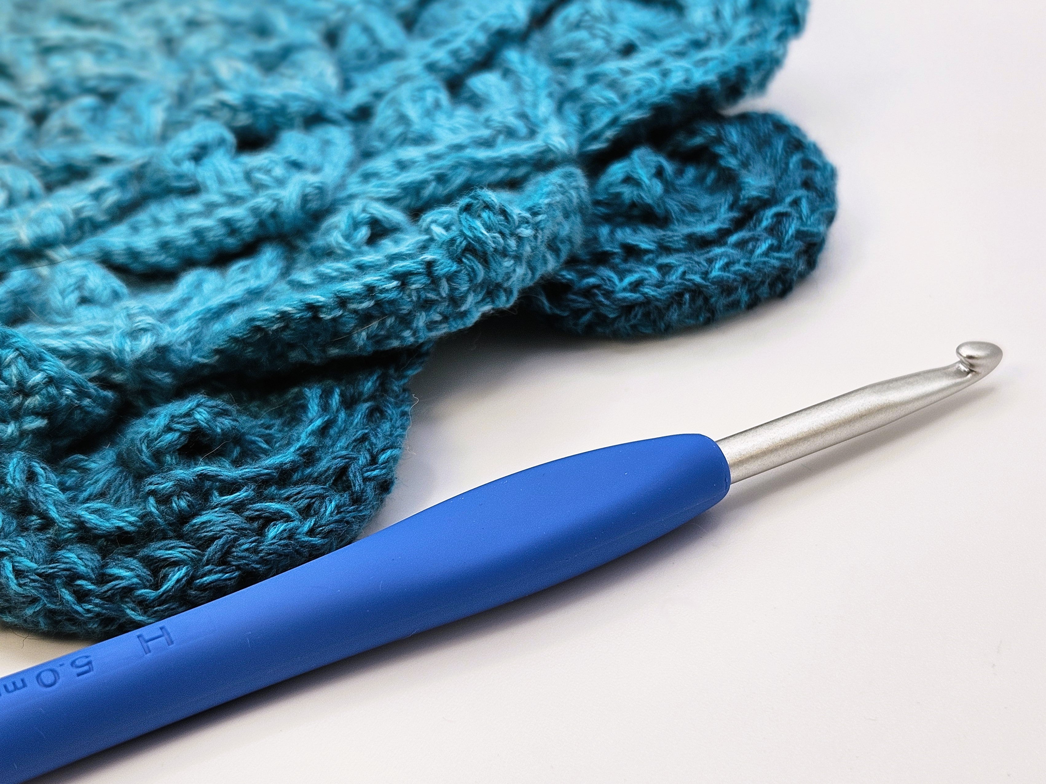 Clover Amour Crochet Hook in size 5.0mm with a blue handle, displayed on a white surface alongside a crocheted teal crochet project.