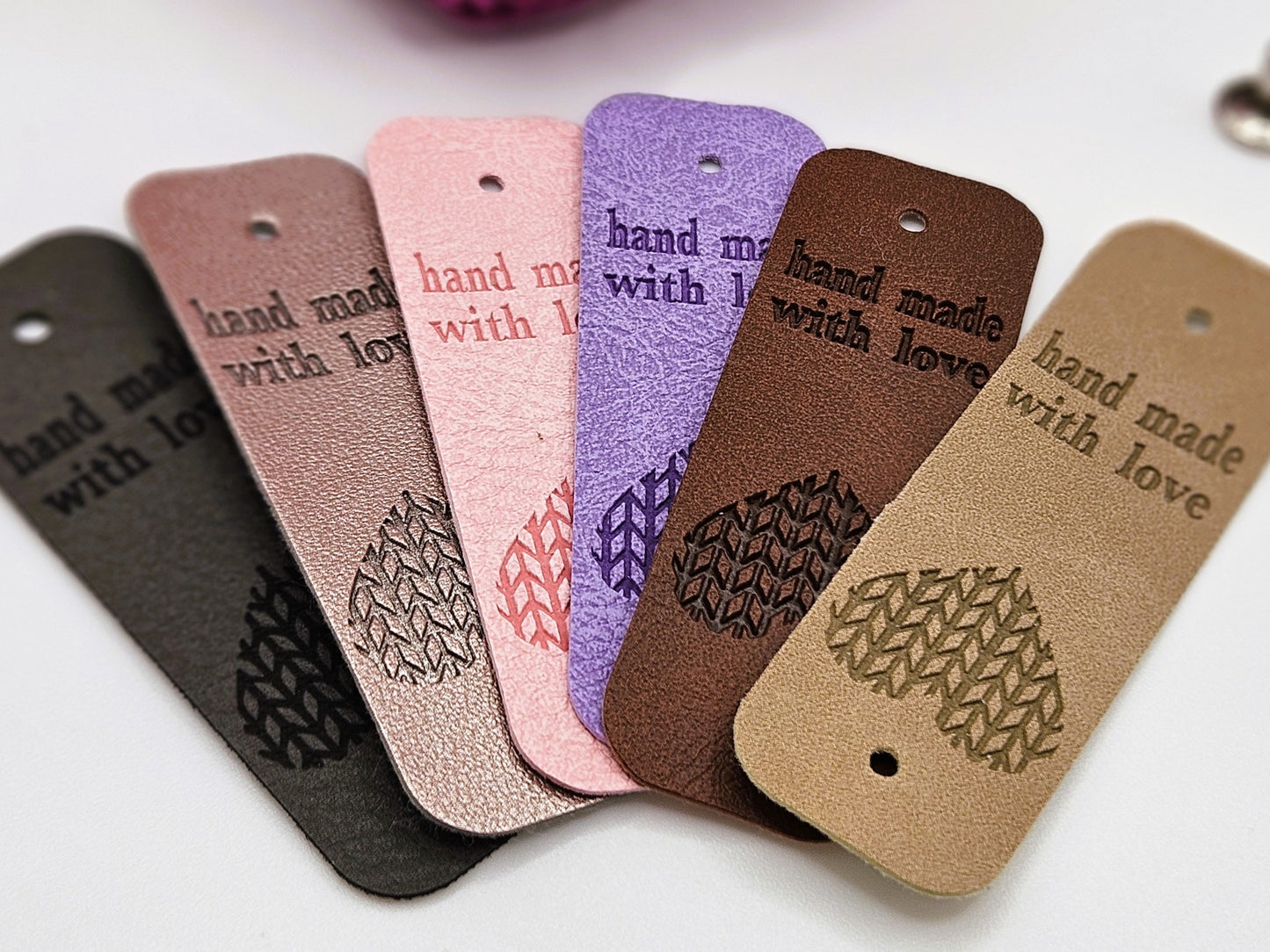 6 faux leather tags for crochet projects with the writing hand made with love on one side and a heart design on the other side. the tags are laid in a fan shape and are in colours dark grey, silver, pink, purple, dark brown and beige.