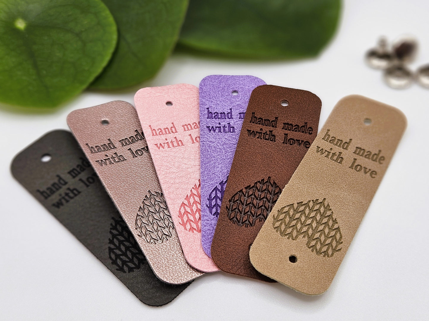 6 hand made with love tags with the writing 'hand made with love' on one end and a heart shaped design on the other end. The hand made tags are laid out in a fan shape. The colours are dark grey, silver, pink, purple, brown and beige.