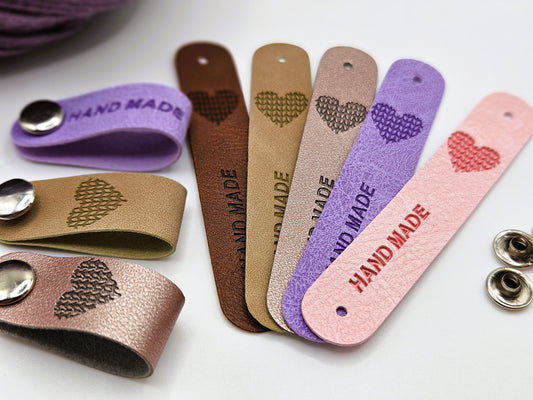 A collection of faux leather hand-made tags in different colours (beige, brown, pink, purple, and silver) with snap-on poppers, scattered around, showing the heart-shaped design and the text hand made.