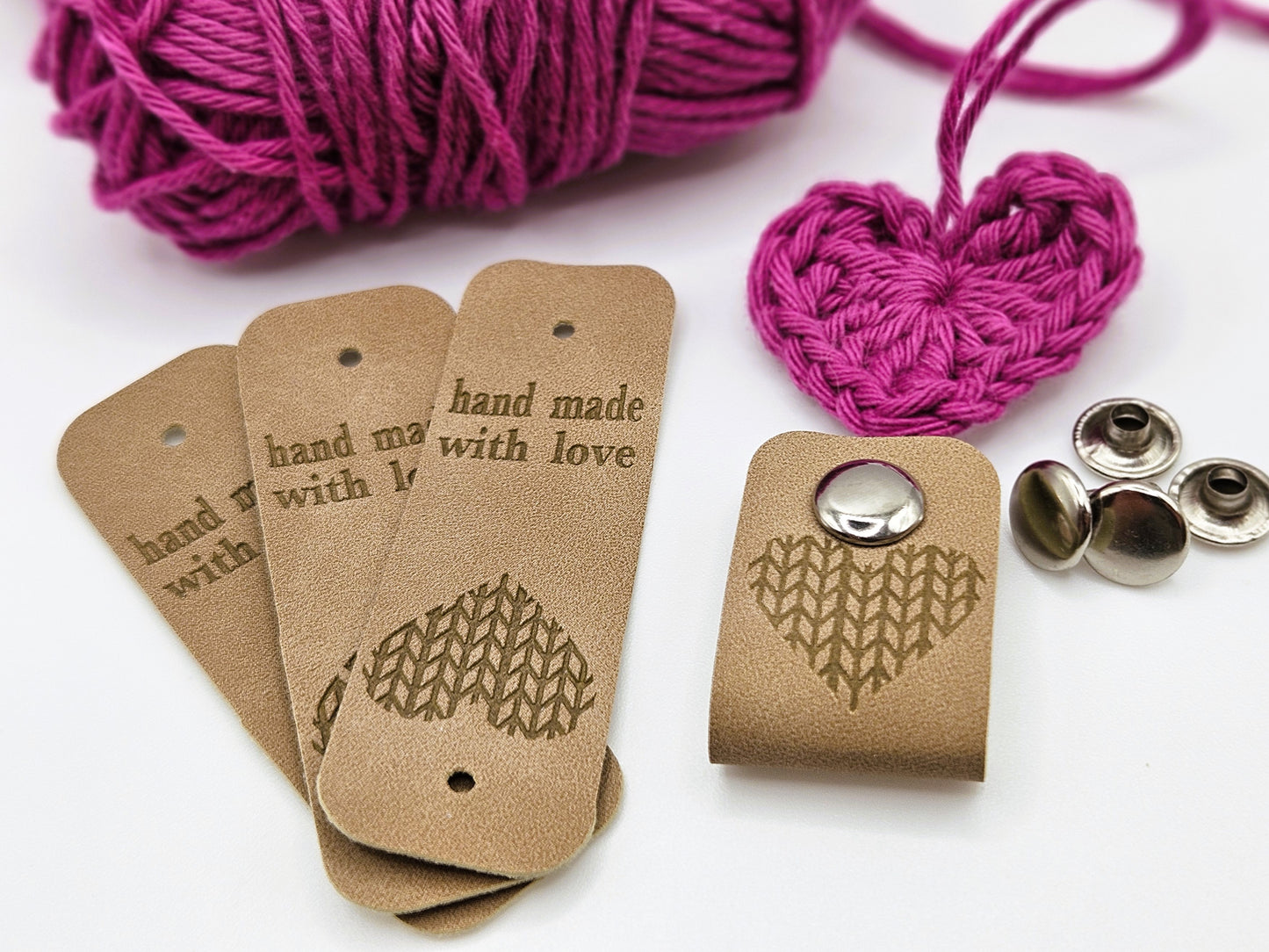 Three beige faux leather hand made with love tags laid in a fan design. To the right is a hand made with love tag folded in half and secured with a metal popper. There is a heart design on the tag. There are four metal poppers in the background.
