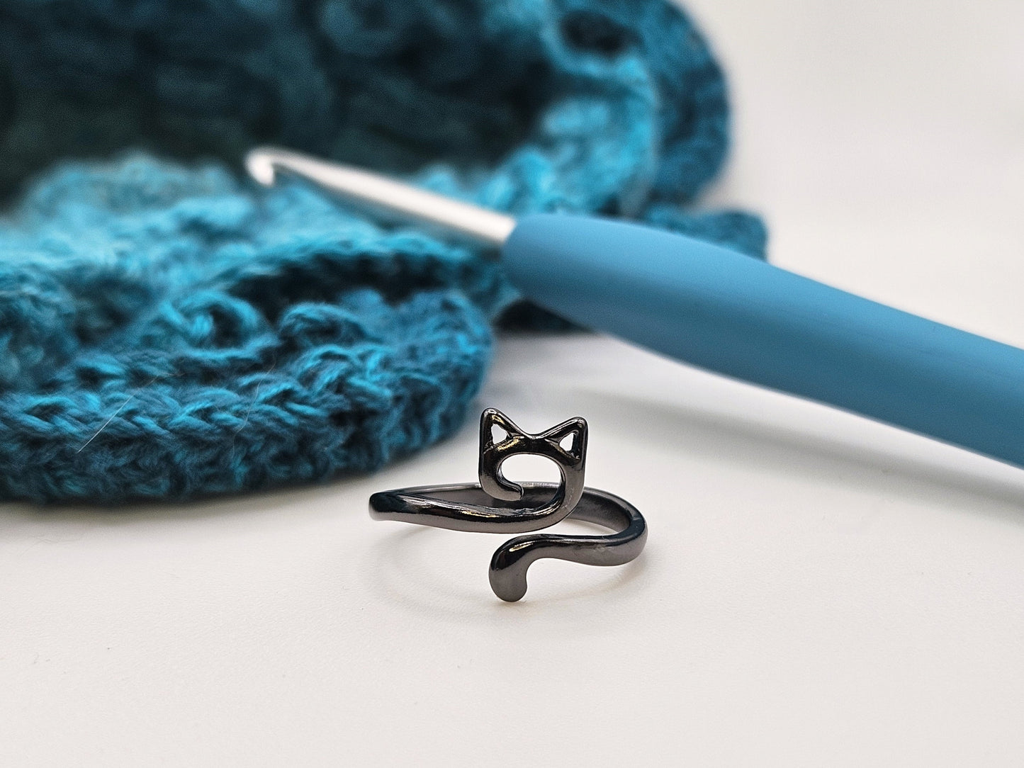 Black cat-shaped crochet tension ring displayed on a white surface, with a blue crochet hook and a crocheted teal project in the background.