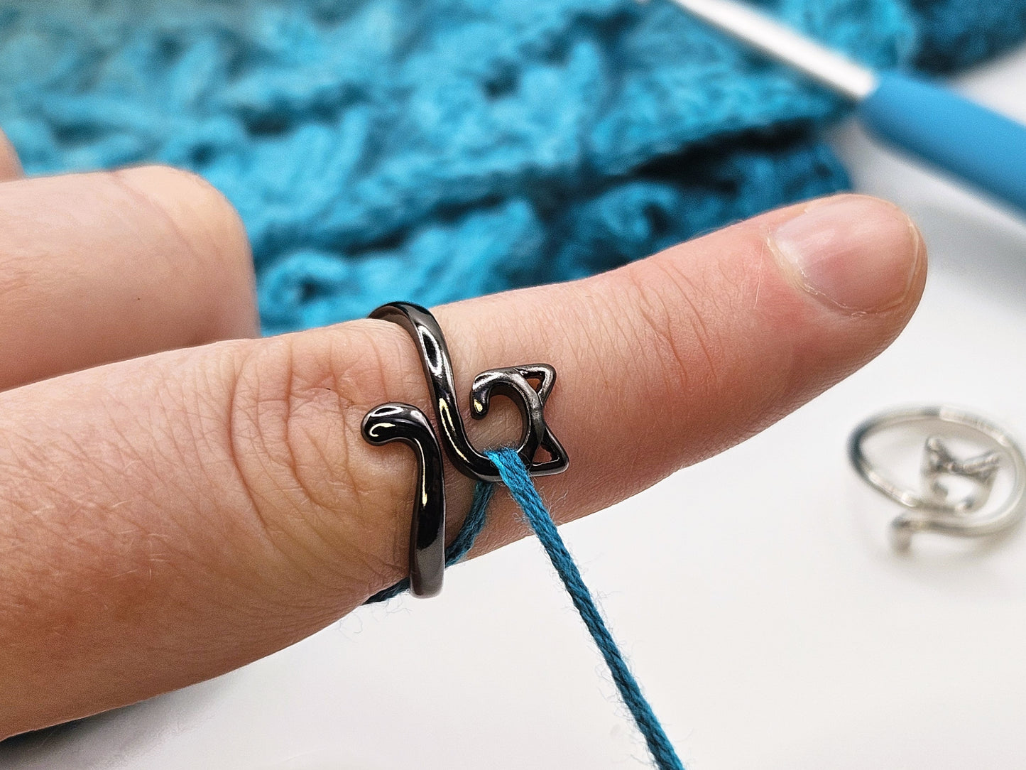 Black cat-shaped crochet tension ring worn on a finger, holding a strand of teal yarn, with a teal crochet project, crochet hook, and a silver crochet ring in the background