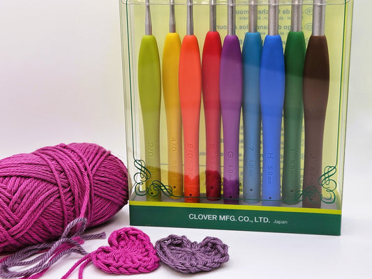 Box of Clover Amour Crochet Hooks in various sizes and colours, displayed alongside a ball of pink yarn and two crocheted hearts.