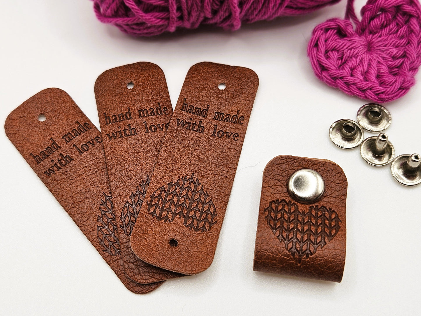 Three brown faux leather hand made with love tags laid in a fan design. To the right is a hand made with love tag folded in half and secured with a metal popper. There is a heart design on the tag. There are four metal poppers in the background.