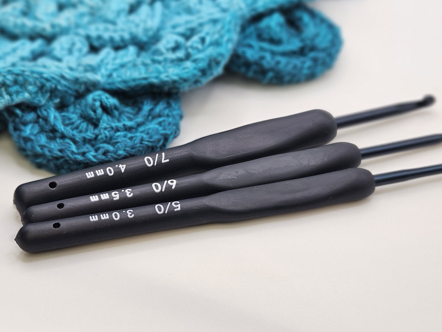 Three soft grip ergonomic crochet hooks with black rubberised handles in sizes 4.0mm, 3.5mm, and 3.0mm, displayed on a white surface with a crocheted teal project in the background.