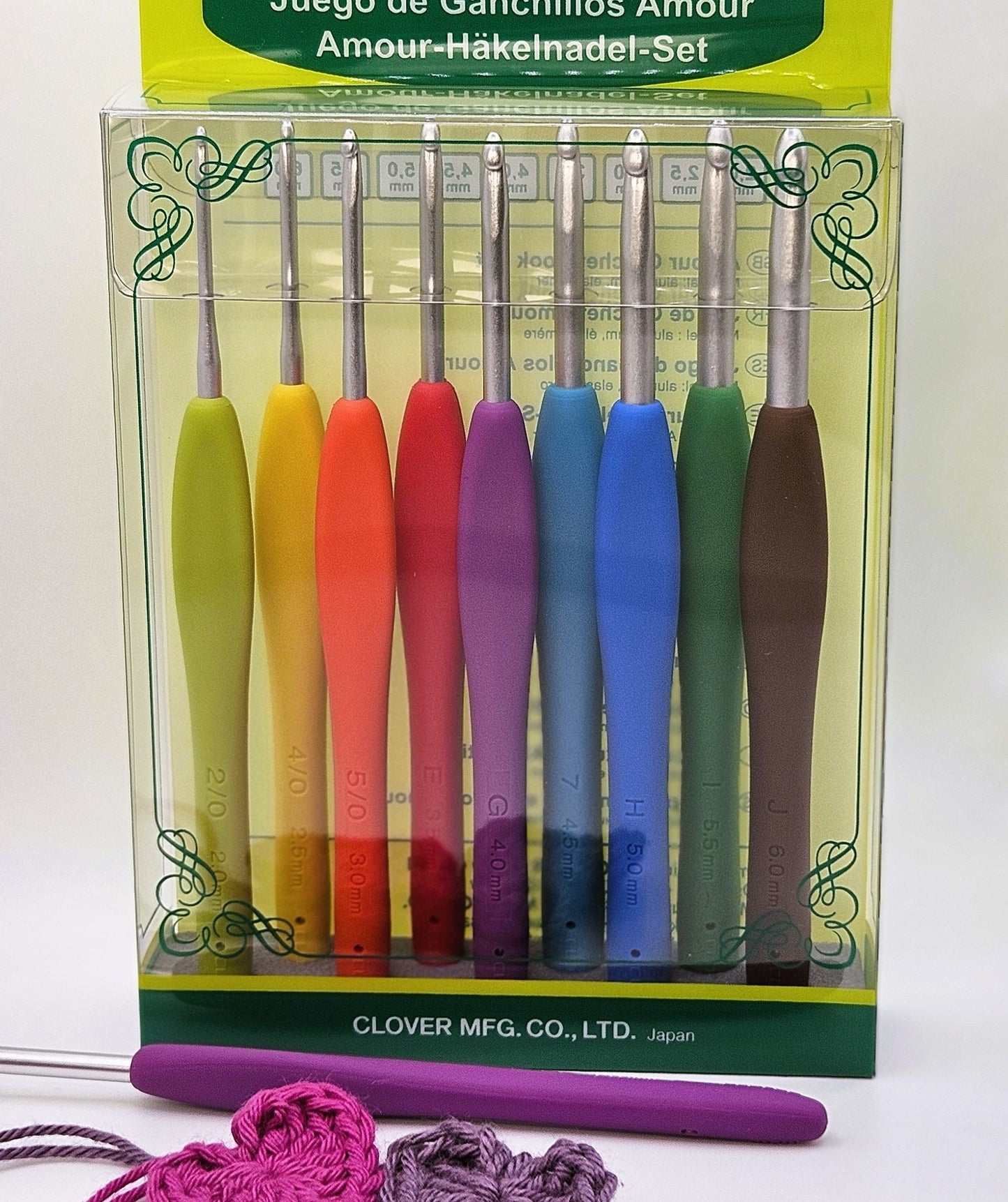 Box of Clover Amour Crochet Hooks in a range of sizes and colours, displayed with a purple-handled crochet hook and two crocheted hearts in pink and purple yarn.