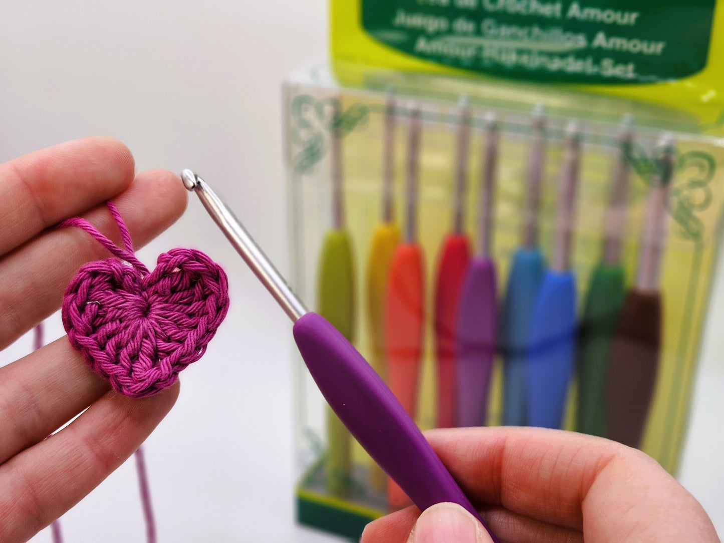 Clover Amour Crochet Hook in use with pink yarn to create a crocheted heart, with the full hook set displayed in the background.