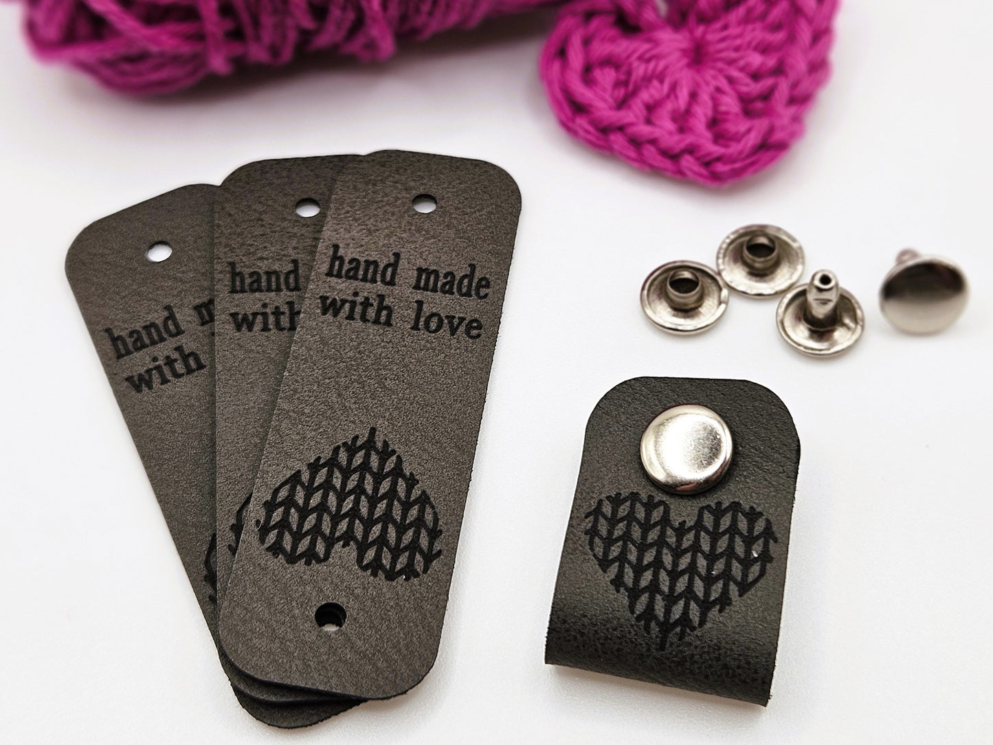 Three dark grey faux leather hand made with love tags laid in a fan design. To the right is a hand made with love tag folded in half and secured with a metal popper. There is a heart design on the tag. There are four metal poppers in the background.