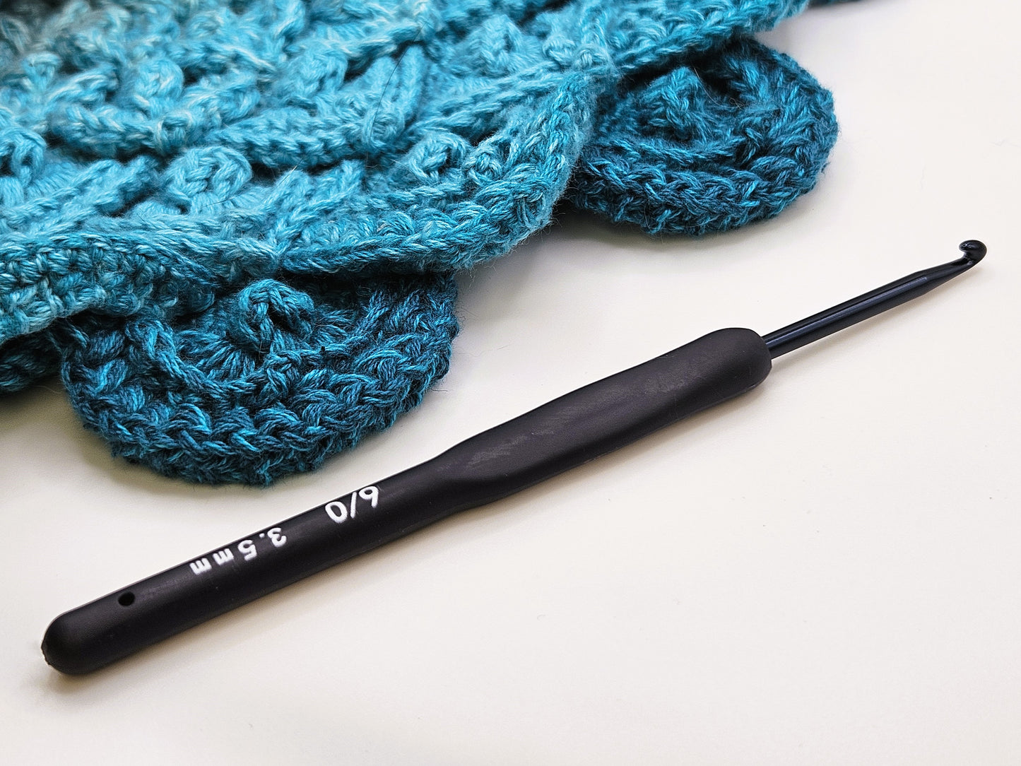 Soft grip ergonomic crochet hook with a black rubberised handle, size 3.5mm, displayed on a white surface next to a crocheted teal project.