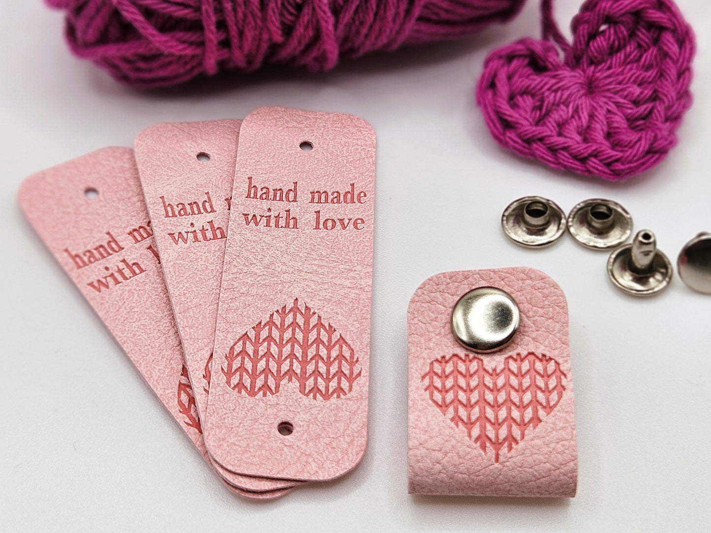Three pink faux leather hand made with love tags laid in a fan design. To the right is a hand made with love tag folded in half and secured with a metal popper. There is a heart design on the tag. There are four metal poppers in the background.