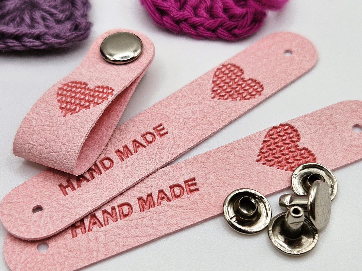 Pink faux leather hand made tag with a snap-on popper, engraved with the word "HAND MADE" and a heart design.