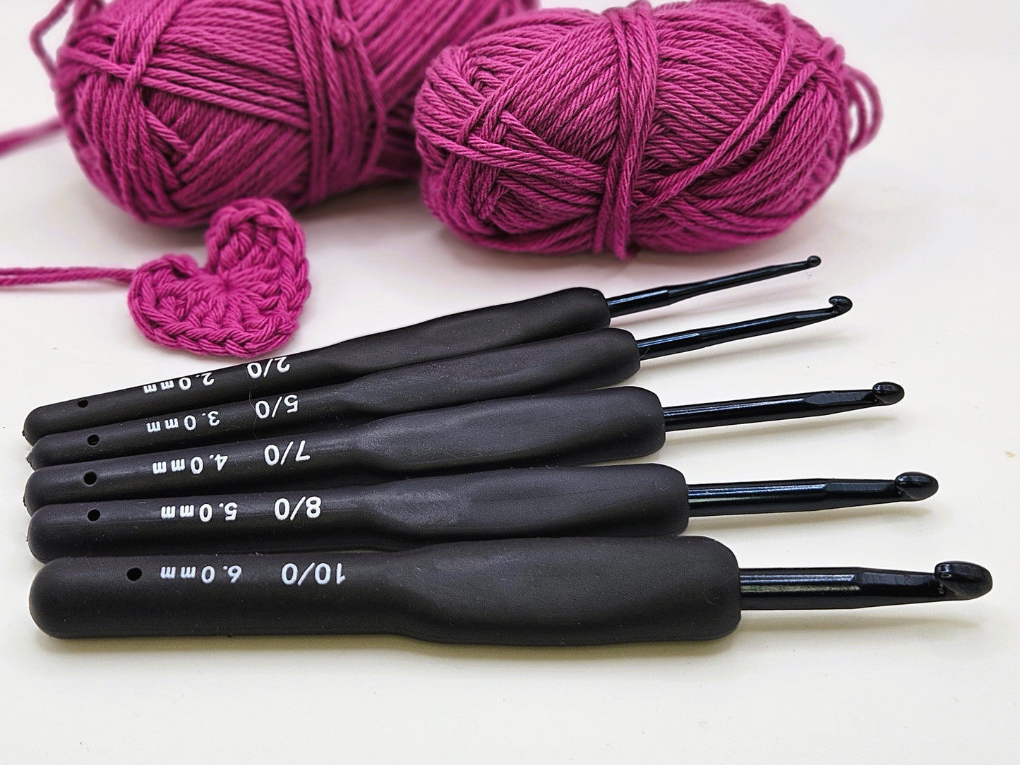 Set of five soft grip ergonomic crochet hooks with black rubberised handles, arranged on a white surface alongside two balls of pink yarn and a crocheted heart.