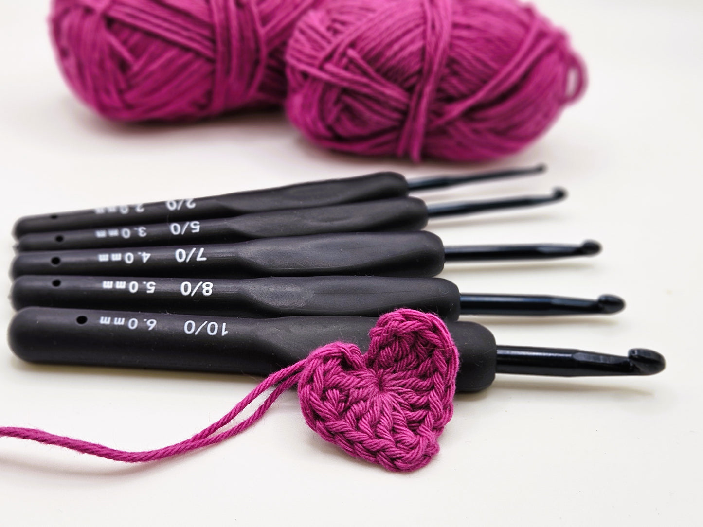 Set of soft grip ergonomic crochet hooks with black rubberised handles, displayed with a crocheted pink heart and two balls of matching pink yarn in the background.