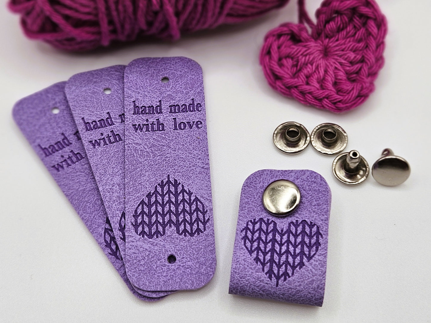 Three purple faux leather hand made with love tags laid in a fan design. To the right is a hand made with love tag folded in half and secured with a metal popper. There is a heart design on the tag. There are four metal poppers in the background.
