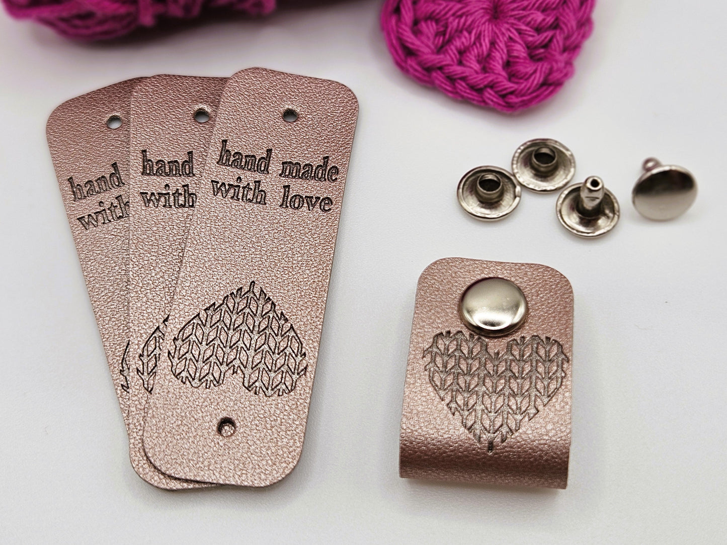 Three silver faux leather hand made with love tags laid in a fan design. To the right is a hand made with love tag folded in half and secured with a metal popper. There is a heart design on the tag. There are four metal poppers in the background.