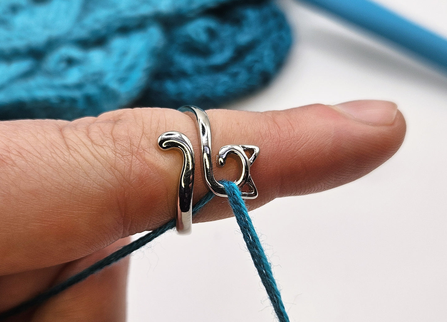 Silver cat-shaped crochet tension ring worn on a finger, holding a strand of teal yarn, with a knitted teal project and crochet hook in the background