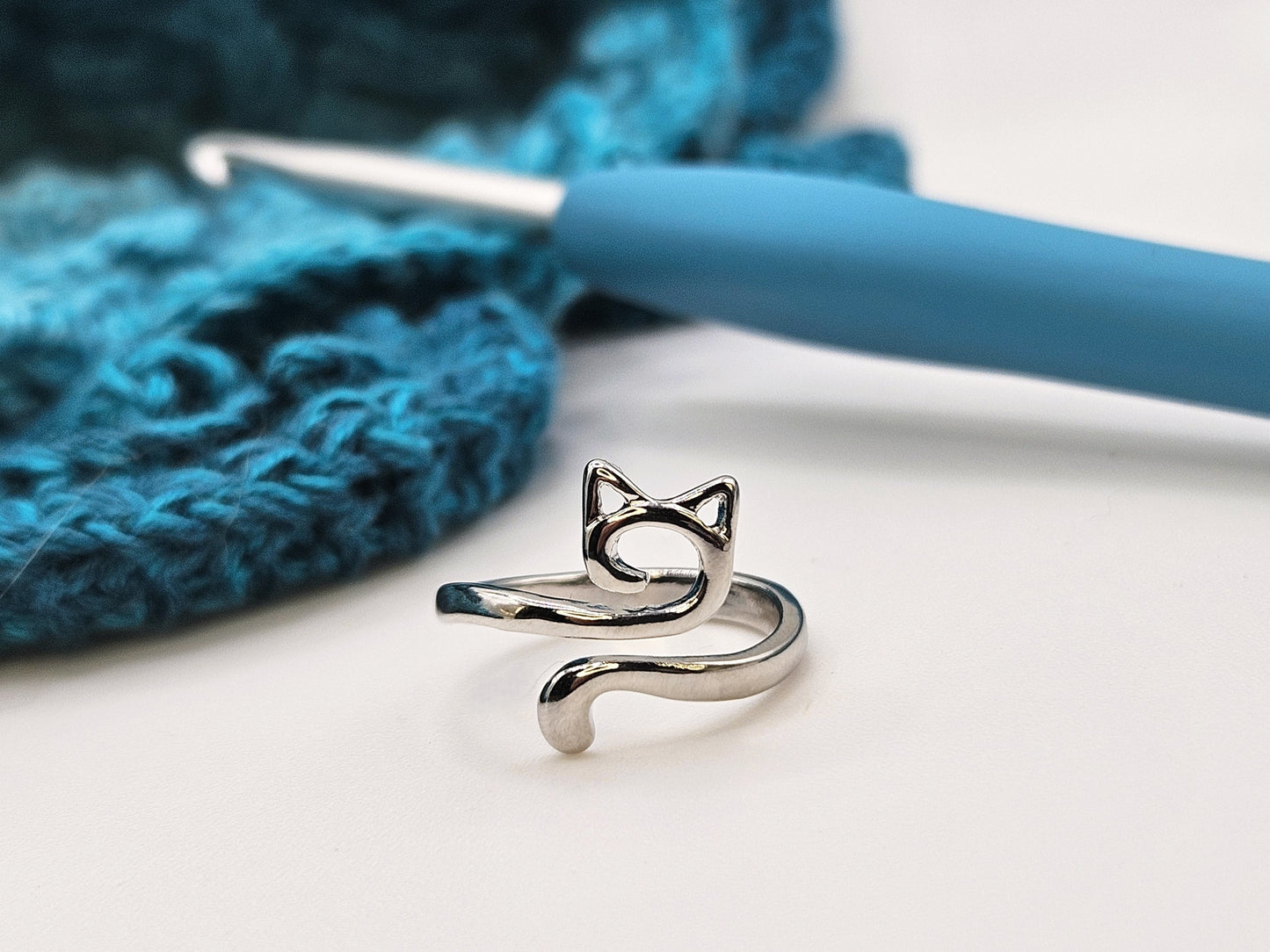Silver cat-shaped crochet tension ring placed on a white surface, with a blue crochet hook and a teal crochet project in the background.