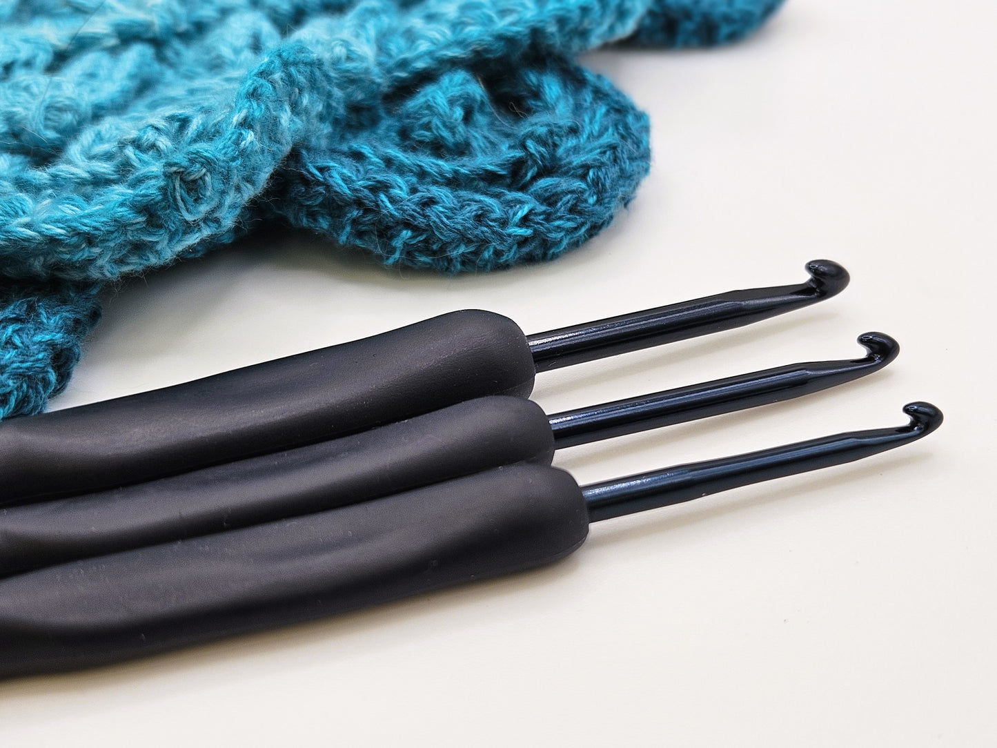 Close-up of three soft grip ergonomic crochet hooks with black rubberised handles, placed on a white surface next to a crocheted teal project.