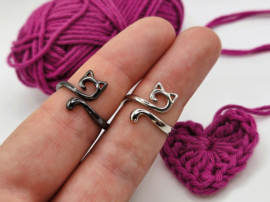 Two cat-shaped crochet tension  rings, one black and one silver, displayed on fingers with a ball of pink yarn and a crocheted heart in the background.