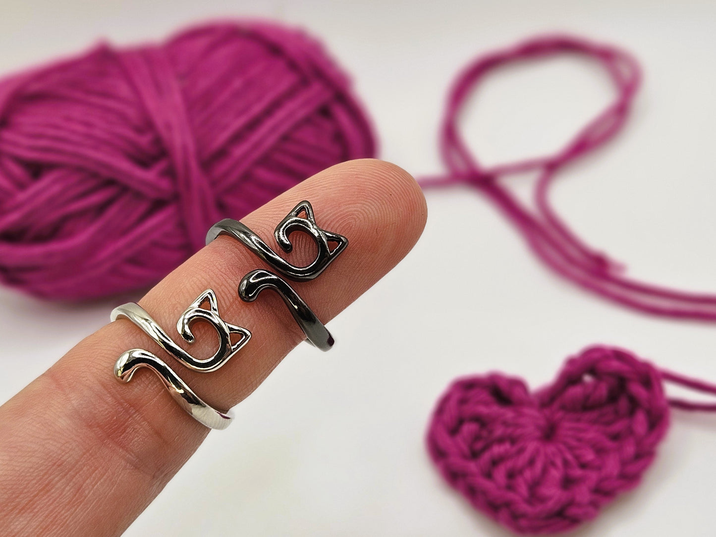 Silver and black cat-shaped crochet tension rings displayed on a finger, with a ball of pink yarn and a crocheted heart in the background.