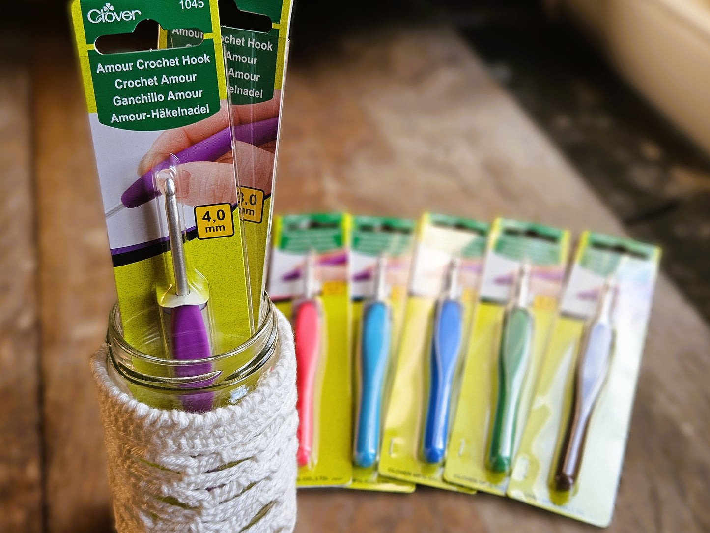 Clover Amour Crochet Hook in size 4.0mm packaged and displayed in a jar wrapped with crocheted white yarn, with other packaged hooks in various sizes arranged in the background.