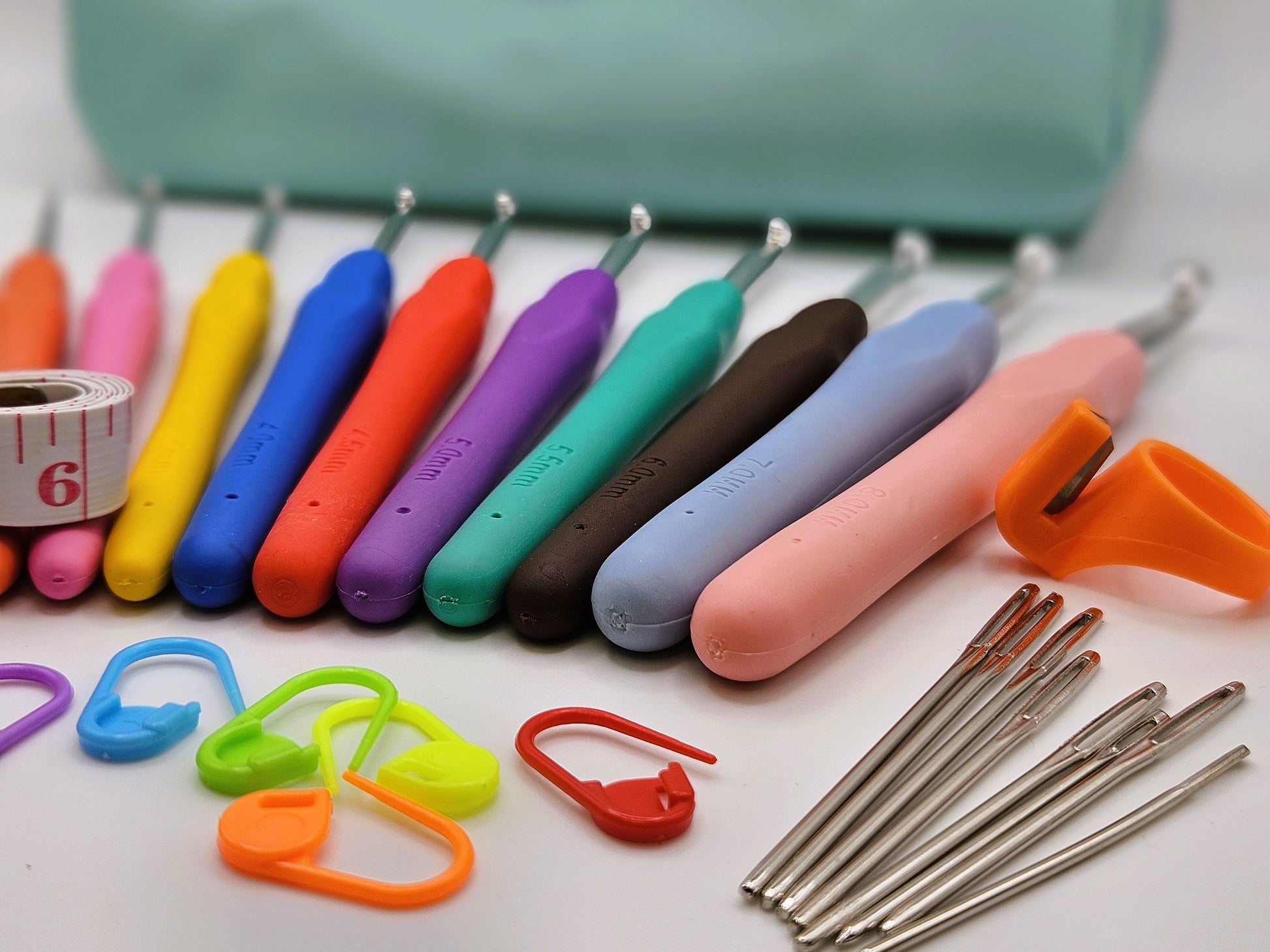 Set of colourful soft grip ergonomic crochet hooks, stitch markers, darning needles, a thread cutter, and a measuring tape arranged neatly on a white surface with a storage case in the background.