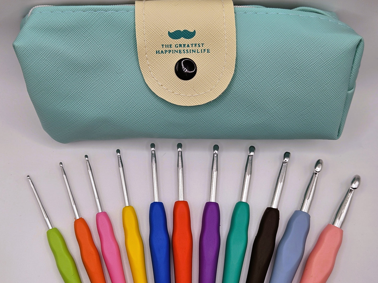 Close-up of colourful soft grip crochet hooks laid out below a teal storage case with a snap closure, featuring the text "The Greatest Happiness in Life."
