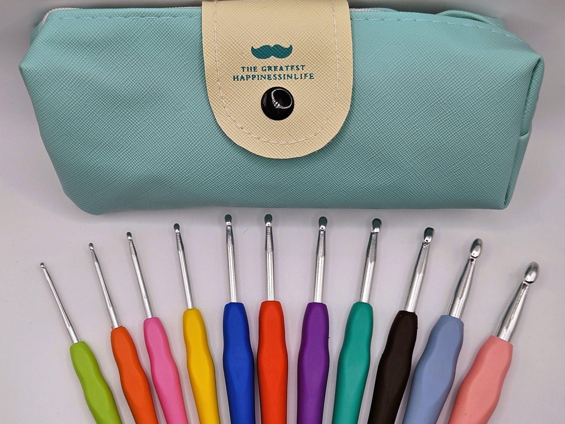 Close-up of colourful soft grip crochet hooks laid out below a teal storage case with a snap closure, featuring the text "The Greatest Happiness in Life."