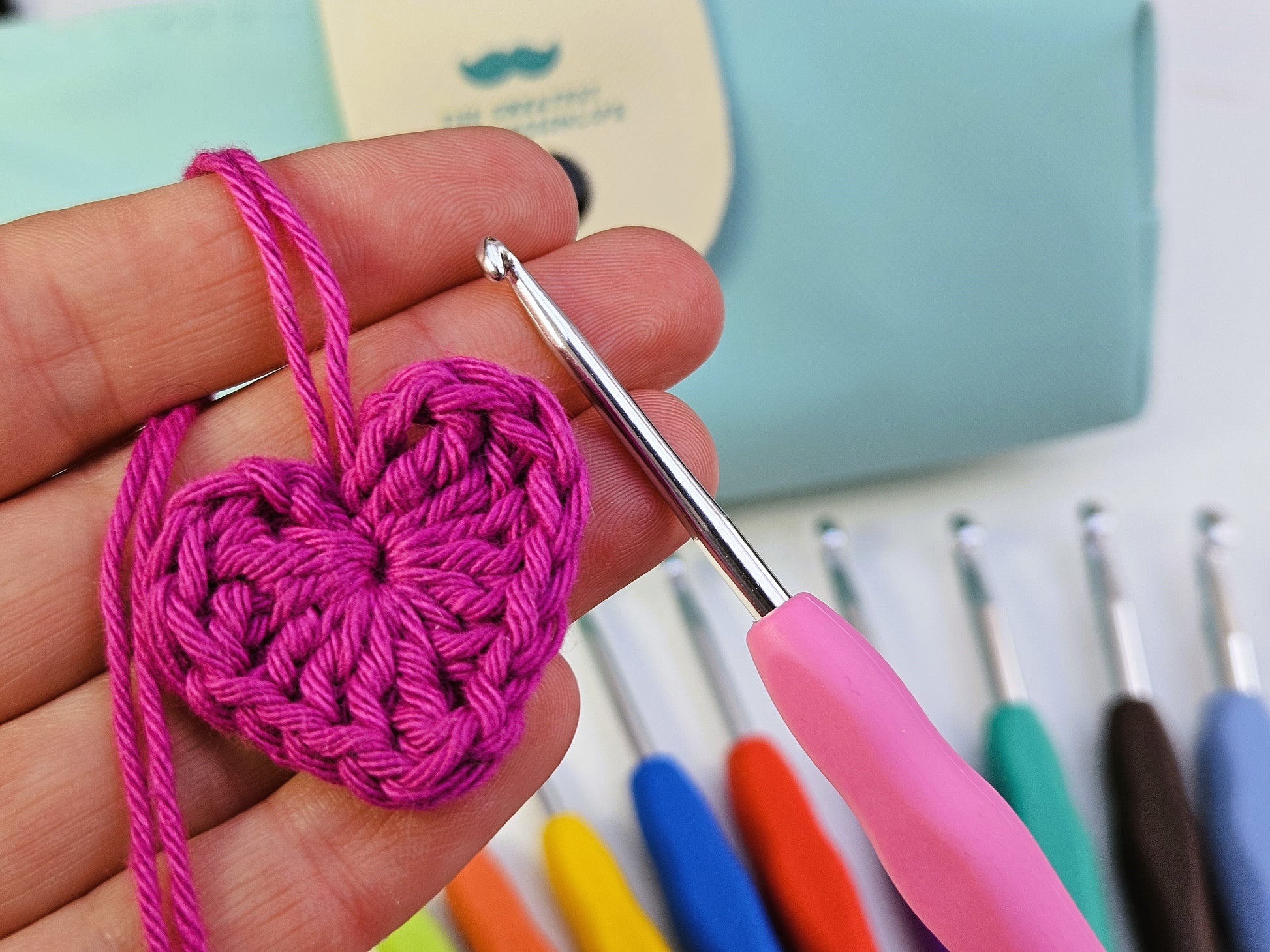 Close-up of a pink soft grip crochet hook in use, creating a crocheted heart with pink yarn, mint green storage case visible in the background.