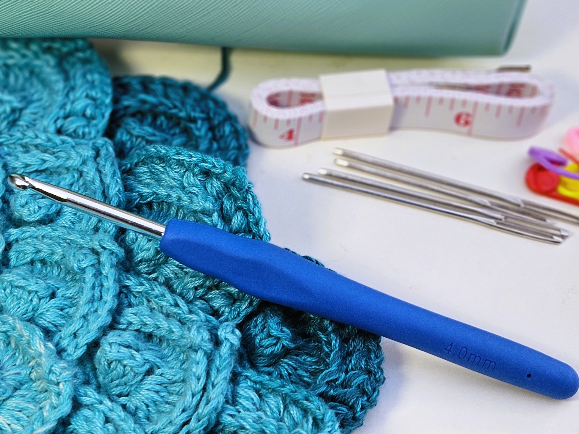 Blue soft grip ergonomic crochet hook displayed on top of a teal crocheted project, with darning needles, stitch markers, and a measuring tape visible in the background.