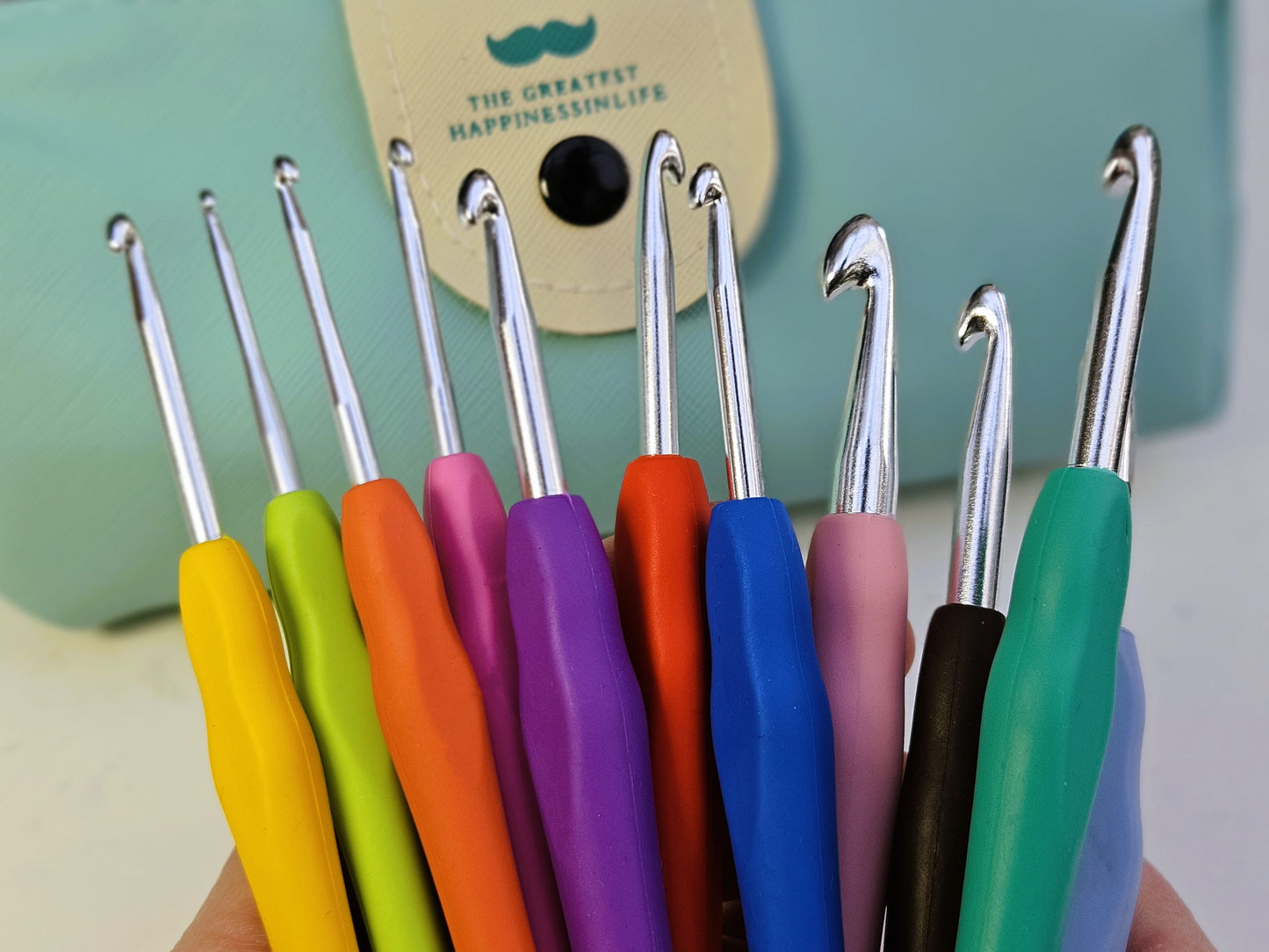 A close-up of the metallic tips of colourful soft grip ergonomic crochet hooks, held in hand with a teal storage case in the background.