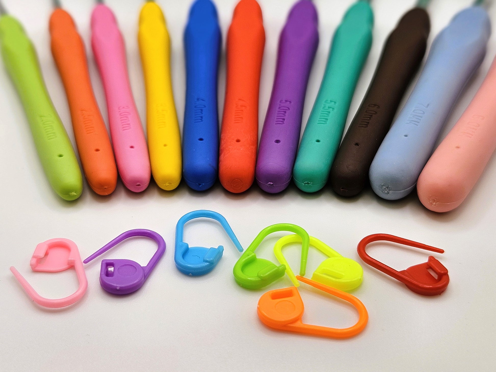 Close-up of colourful soft grip ergonomic crochet hooks lined up, with brightly coloured stitch markers scattered below.